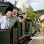 Ausflug mit der Dampflok im Waldviertel - Ferienwohnungen Gutenbrunn Ein Paar winkt fröhlich aus einer historischen Dampflokomotive, ideal für einen familienfreundlichen Ausflug nahe Ferienwohnungen Anna & Herbert Gutenbrunn.