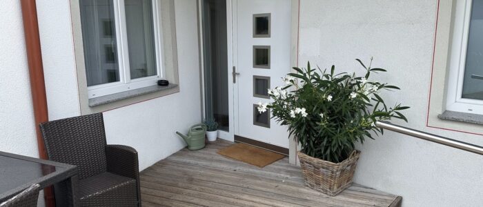vuTuMcqBTY68s14isTovXQ White front door with four square glass inserts on a wooden porch, with a window to the left and a large potted plant in a wicker basket to the right.