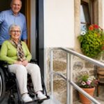 ausstattung-fur-barrierefreie-ferienunterkunfte-checkliste-1792453789 - Ferienwohnungen Gutenbrunn Elderly woman in a wheelchair at a doorway ramp, with a smiling man behind her and potted flowers outside.