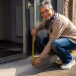 barrierefreie-unterkunft-turen-und-stufen-prufen-1791792920 - Ferienwohnungen Gutenbrunn Woman crouching by an open front door, measuring the threshold with a tape measure outdoors.