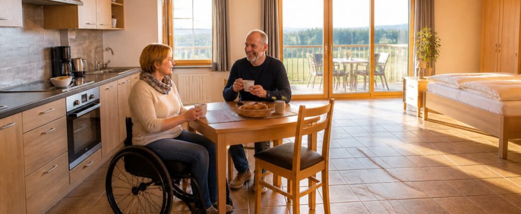 accessible-accommodation-kitchen-living-room-bedroom-1791611148-50 - Gutenbrunn Vacation Rentals A couple sharing coffee at a wooden table in a bright, open-concept kitchen; a woman in a wheelchair is on the left, and a man is smiling across the table.