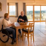barrierefreie-unterkunft-wohnkuche-schlafzimmer-1791611148-50 - Ferienwohnungen Gutenbrunn Couple sharing coffee at a wooden table in a bright open-plan kitchen, a woman in a wheelchair on the left and a man smiling across the table.