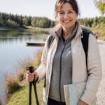 akadálymentes túraútvonalak a tó körül – keresés és tervezés – 1791848940 - Gutenbrunn nyaralók Egy mosolygó nő túrabotokkal túrázik egy tó partján, kezében térképpel és hátizsákkal, egy napsütéses napon egy kavicsos ösvényen; a háttérben az ösvényen egy kerekesszékes személy látható.