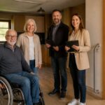 barrierefreiheit-in-ferienwohnungen-sicher-prufen-1790343217-3 - Ferienwohnungen Gutenbrunn Four people stand together inside a home: an older man in a wheelchair, an older woman, and two professionals with clipboards, smiling at the camera.
