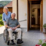 checklist-for-accessible-vacation-rentals-for-seniors-1791348562 - Gutenbrunn Vacation Rentals An elderly man in a wheelchair being pushed by a smiling caregiver outside a home with an open door and a ramp.