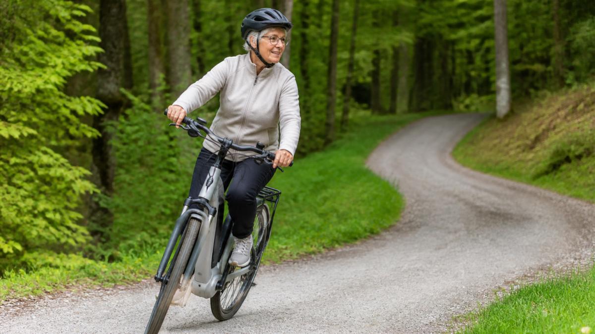 Eine ältere Person sitzt auf einem E-Bike auf einem ruhigen Waldweg, trägt einen Helm und führt eine Kurvenfahrt durch, mit Bäumen und natürlicher Waldlandschaft im Hintergrund