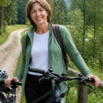 familienfreundliche-radwege-im-waldviertel-7-top-routen-1791935345 - Ferienwohnungen Gutenbrunn Smiling woman in a green jacket with a backpack stands beside a bicycle on a forest dirt path, helmet in tow and a stream nearby.