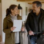 family-accommodations-checklist-amenities-in-the-Waldviertel-1791891518 - Gutenbrunn Vacation Rentals A couple indoors; the woman is holding a checklist and smiling at her partner next to a baby stroller in a bright, homey setting.