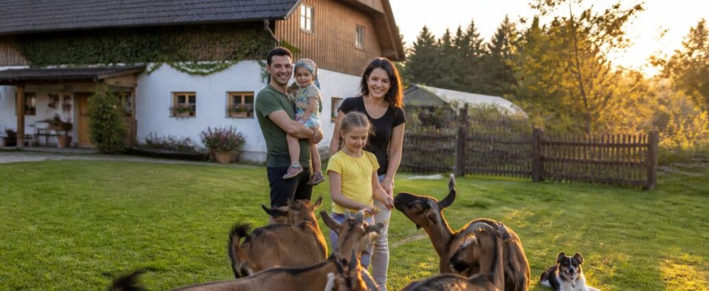 Family-Vacation-on-a-Farm-in-the-Waldviertel-Guide-2026-1791683407 - Gutenbrunn Vacation Rentals A family outside at sunset in a grassy yard; the parents and their two children are interacting with several goats while a dog rests nearby, with a barn behind them.