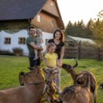 familienurlaub-am-bauernhof-waldviertel-leitfaden-2026-1791683407 - Ferienwohnungen Gutenbrunn Family outside at sunset in a grassy yard, parents with two kids interact with several goats while a dog rests nearby, a barn behind them.