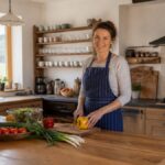 Find-a-Vacation-Rental-with-a-Large-Kitchen-for-Self-Catering-1791176934-1 - Vacation Rentals in Gutenbrunn A woman wearing a striped apron is chopping a yellow bell pepper at a wooden kitchen island, surrounded by fresh vegetables.