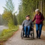 na-invalidnom-vozíku-v-Lesnom kraji-bezbariérové-výlety-1792286525 - Ubytovanie v Gutenbrunne Starší muž na invalidnom vozíku, ktorého tlačí žena po lesnej cestičke vedľa pokojného jazera.